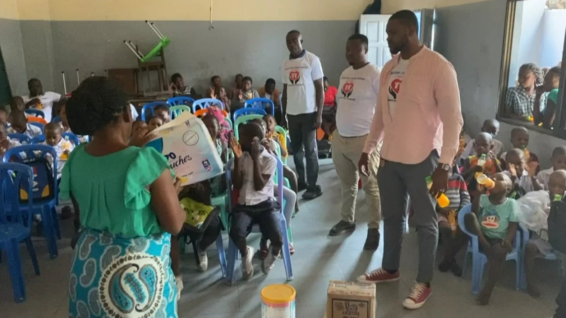 Distribution de dons à des enfants assis dans une salle, en présence de membres de la FOLOK